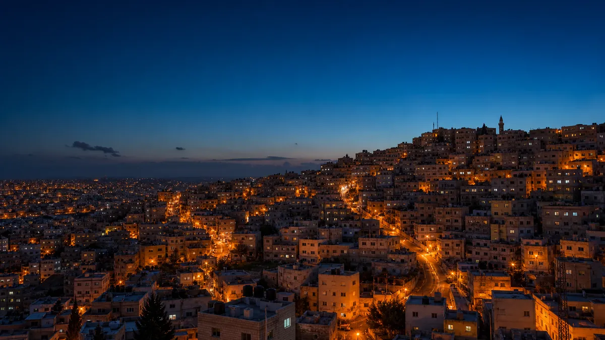 A dense hillside city in the MENA region at dusk, with lit streets running between low-rise buildings.