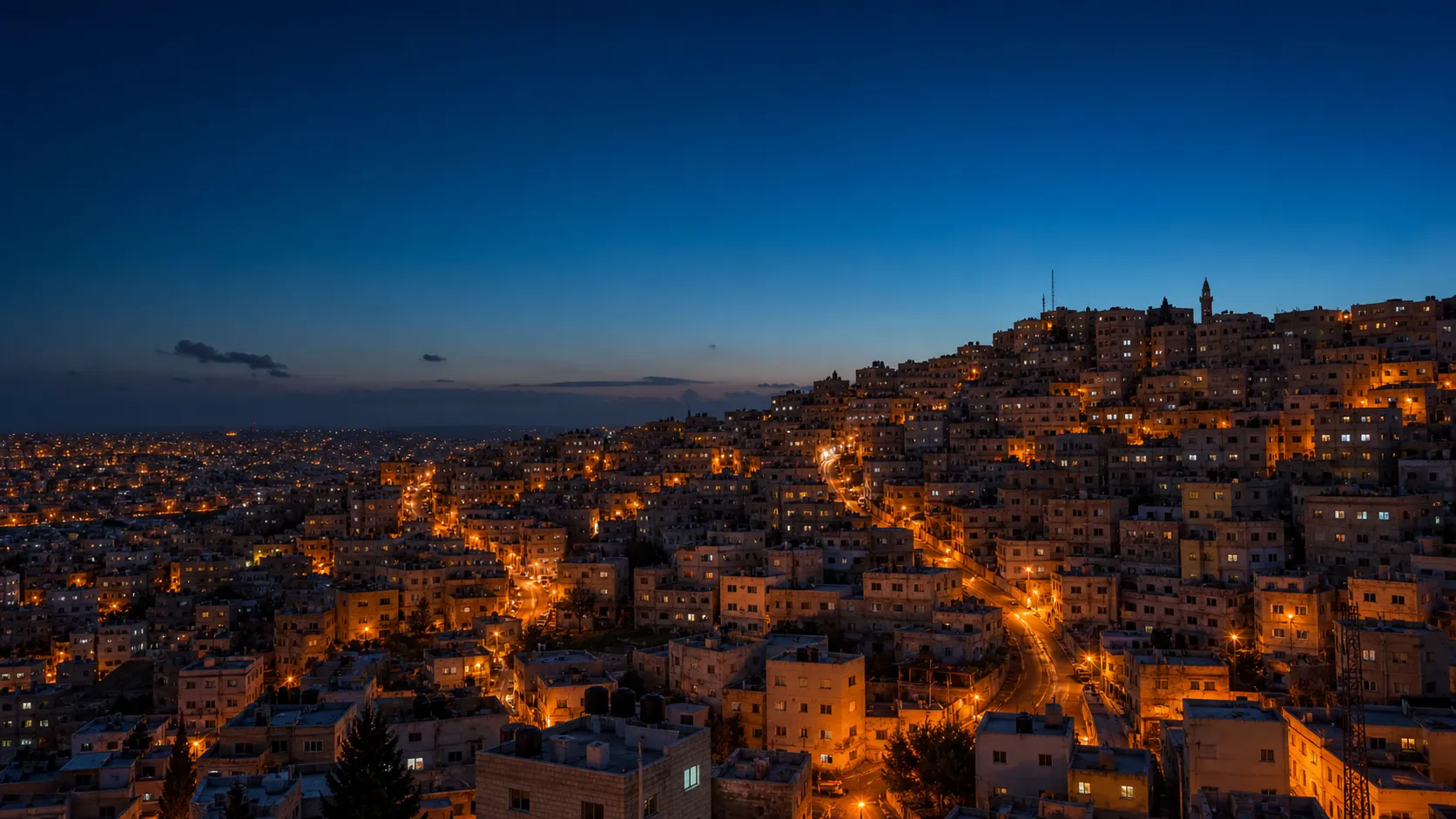 A dense hillside city in the MENA region at dusk, with lit streets running between low-rise buildings.